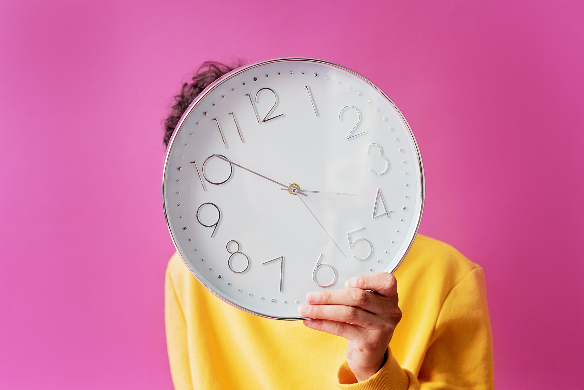 A person in a yellow shirt holds a large wall clock against a vibrant pink background.