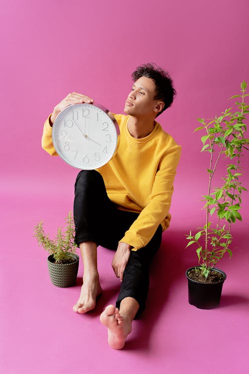 Photo of a Man Holding A Wall Clock Beside Green Plants With Purple Background