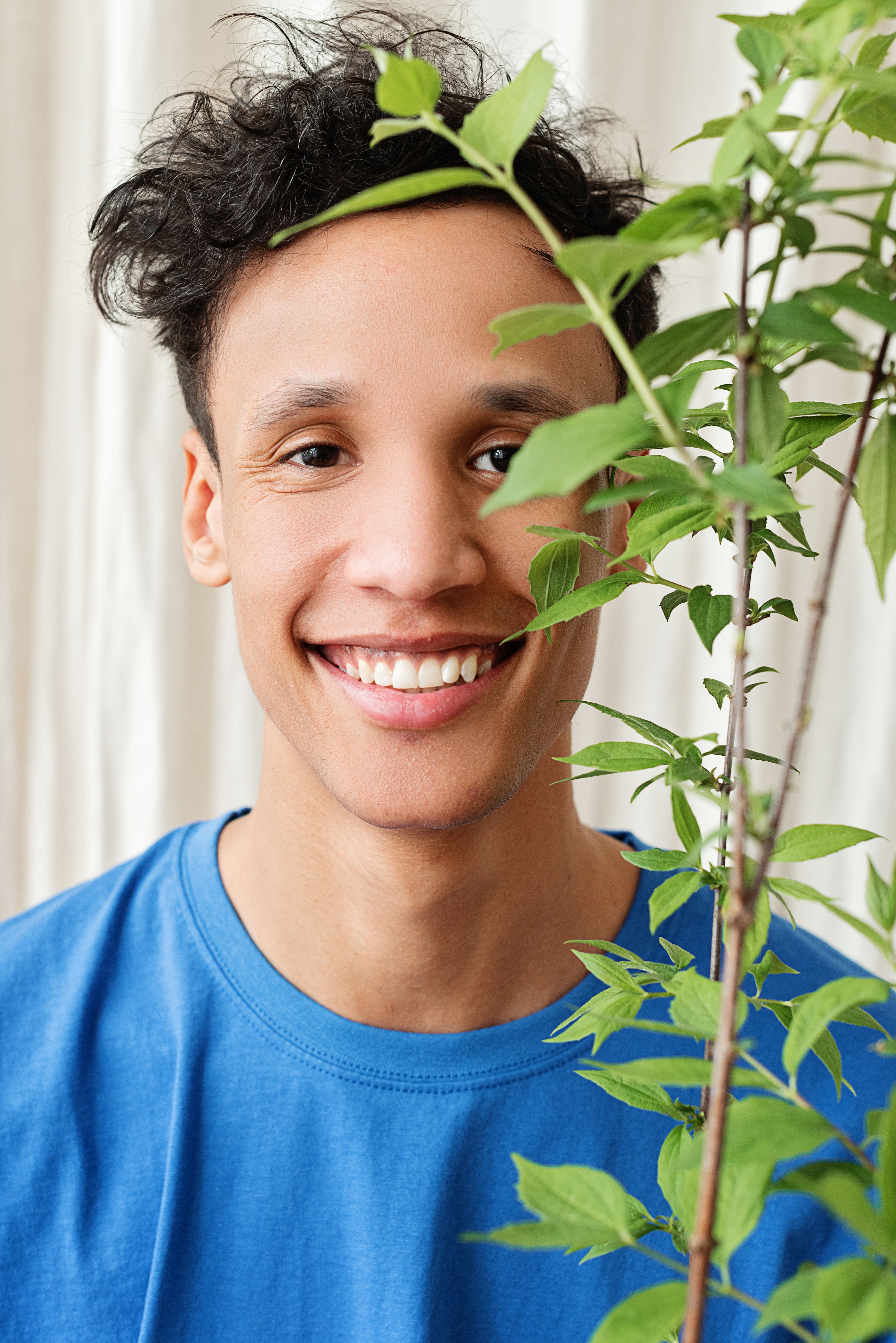 Bright portrait of a smiling young man in a blue shirt, standing near a green plant indoors.