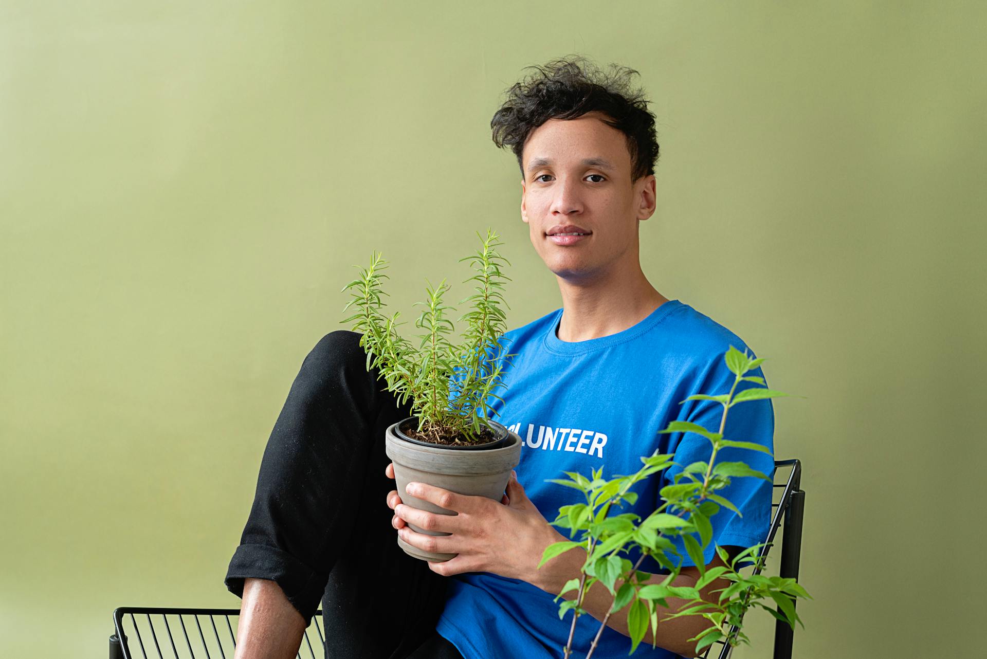 A young man in a blue volunteer shirt holds a potted plant against a green background.