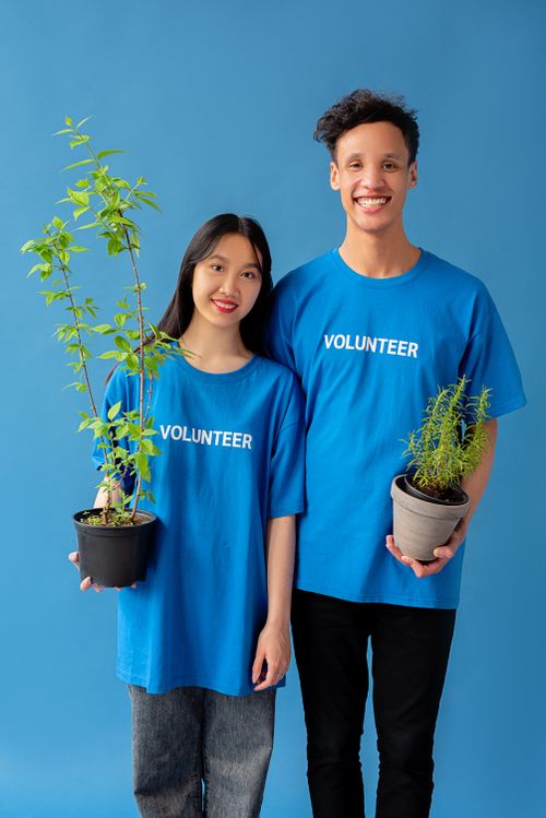 Free Two smiling volunteers in blue shirts hold potted plants against a blue background. Stock Photo