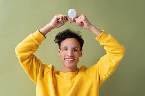 Free A cheerful young adult holds a light bulb overhead against a green background. Stock Photo