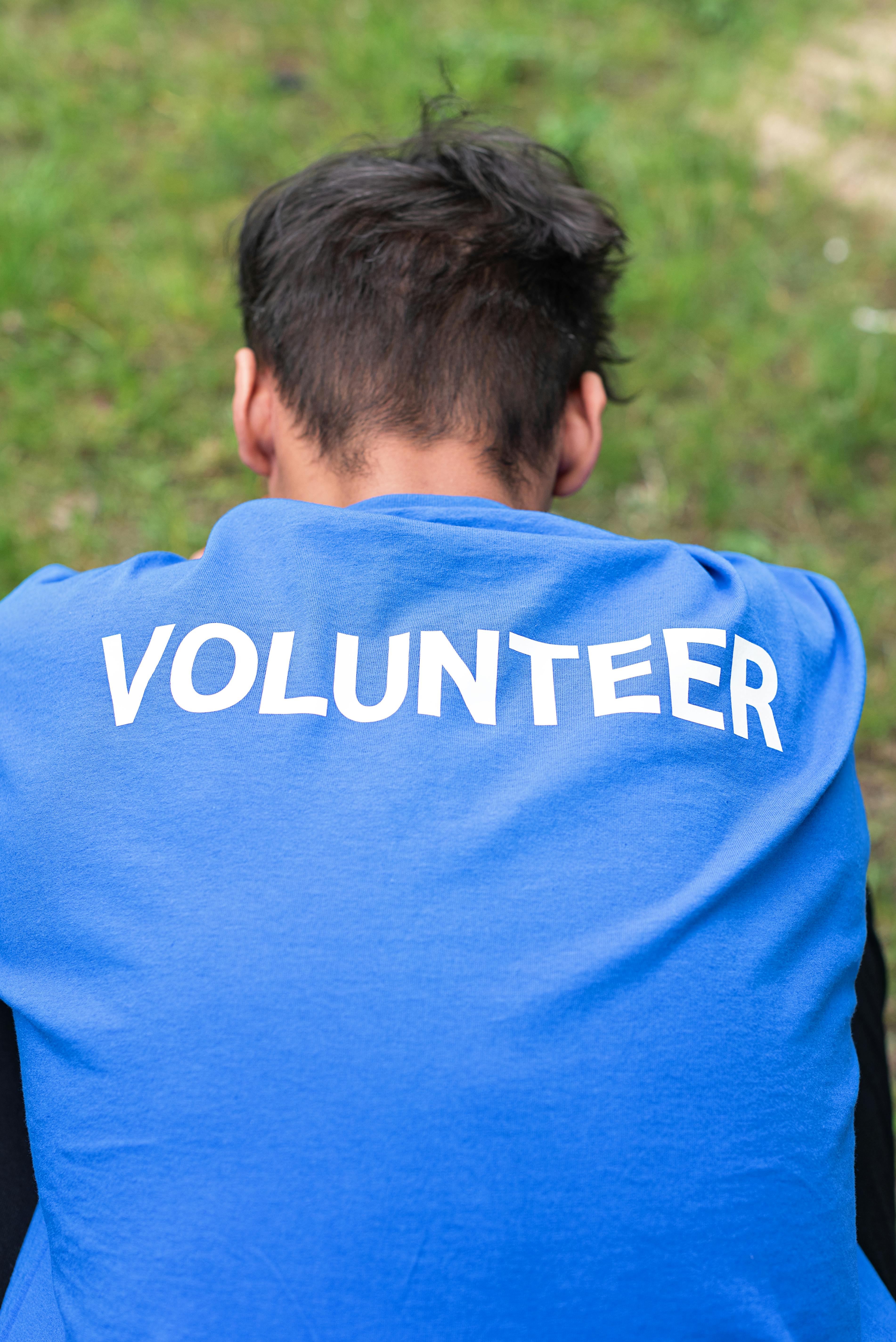 Man wearing a blue volunteer shirt sits on grass, photographed from behind.