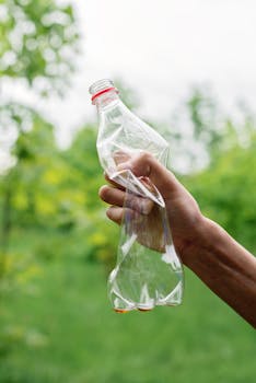 A close-up of a hand holding a crumpled plastic bottle against a green outdoor background.