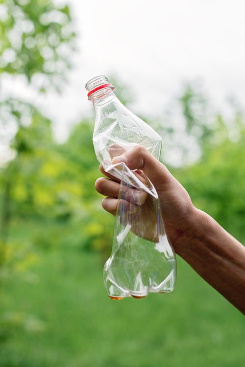 Free A close-up of a hand holding a crumpled plastic bottle against a green outdoor background. Stock Photo