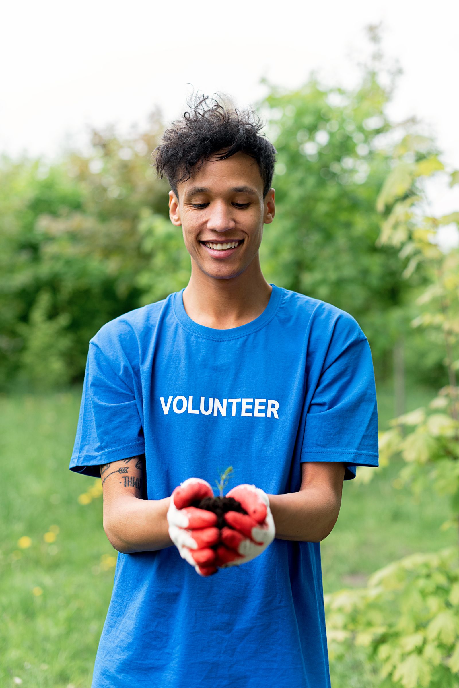 A smiling young volunteer in a blue T-shirt and red gloves holding a seedling in cupped hands