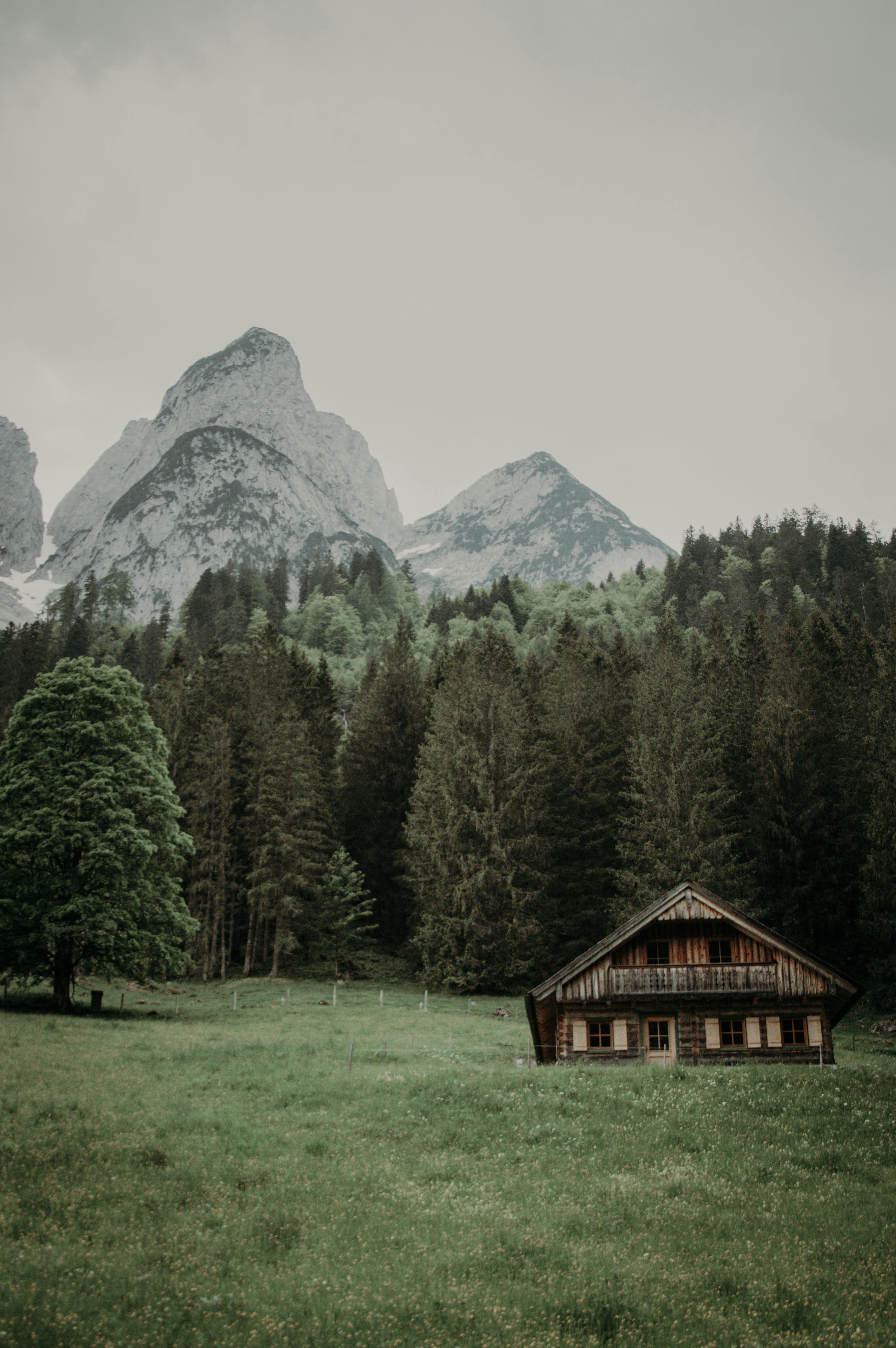 A rustic wooden cabin sits calmly in a lush valley with towering alpine mountains in the background.