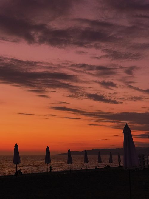 Free A tranquil beach scene at sunset with silhouettes of umbrellas and a vibrant orange sky. Stock Photo