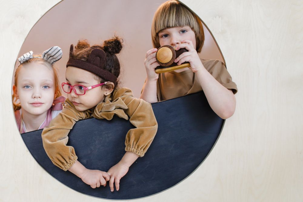 Three smiling children peeking happily through a round wooden window frame