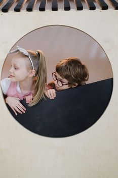 Two kids playing peekaboo through circular window indoors.
