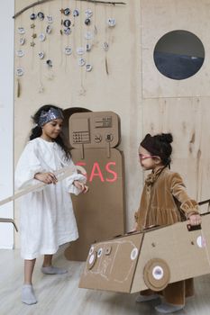 Two young girls engage in imaginative play with a cardboard gas station indoors.