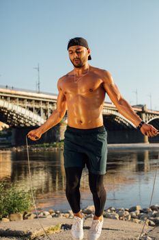 Muscular man skipping rope by river under clear skies, showcasing fitness.