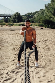 Fit adult man training with battle ropes on a sandy beach outdoors.