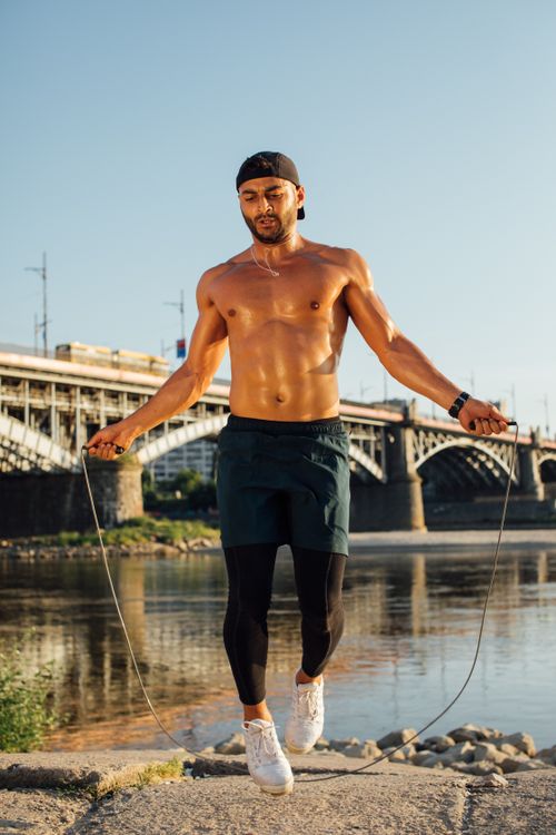 Free Fit man jump roping near a river during a sunny day, showcasing outdoor fitness. Stock Photo