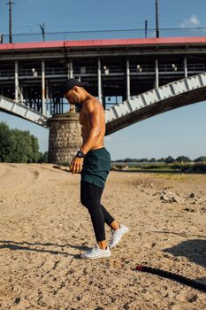 A shirtless man exercising on a sandy beach near a bridge with fitness equipment.
