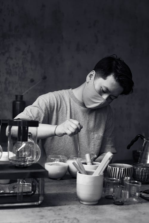 Grayscale Photo of a Man Wearing Face Mask using a Whisk on a Glass Cup