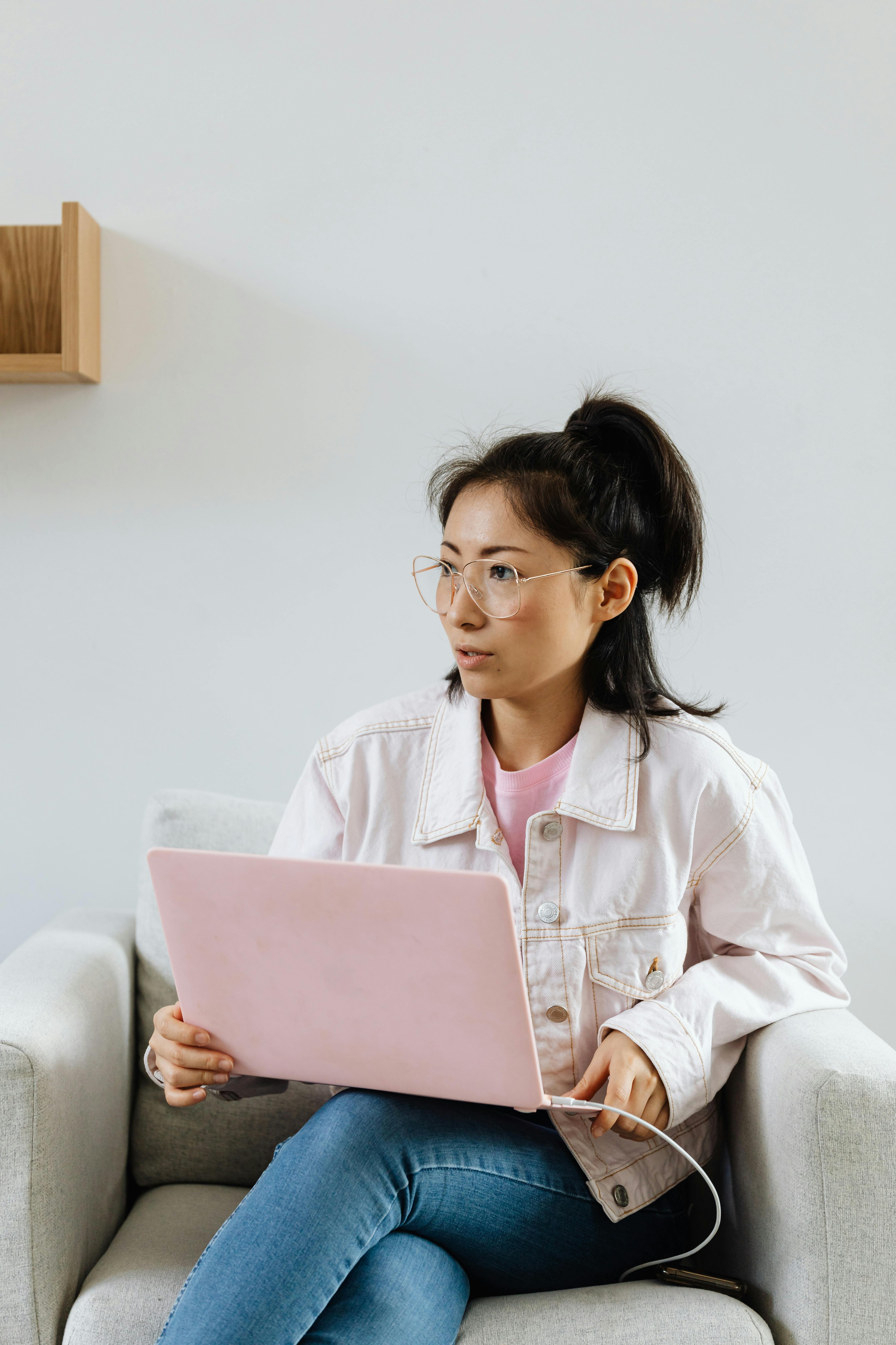 Asian woman sitting on sofa, using laptop; modern lifestyle indoors.