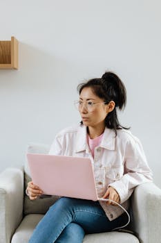 Asian woman sitting on sofa, using laptop; modern lifestyle indoors.