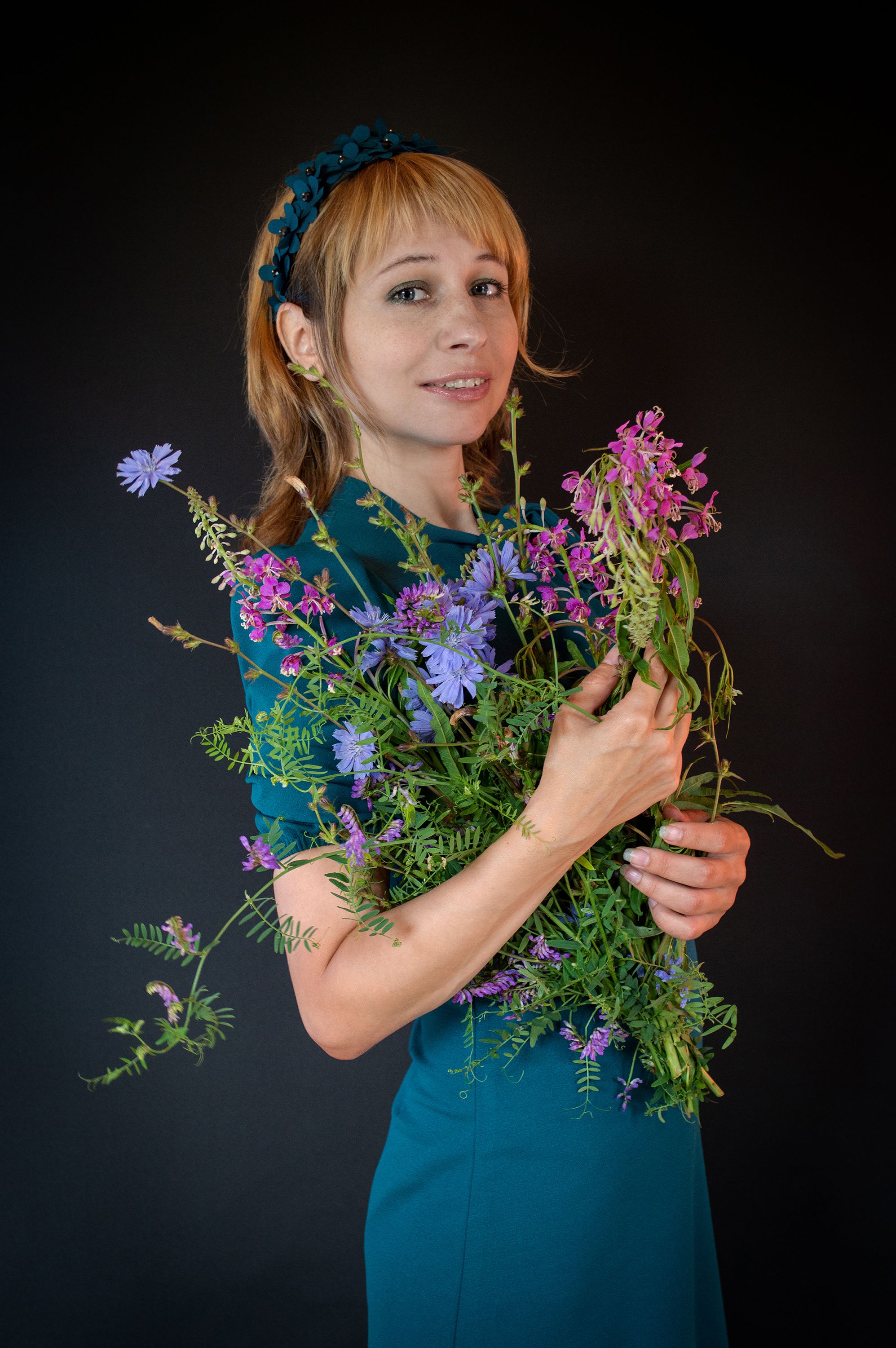 A woman in a blue dress with a floral headband poses with a bouquet against a black background.