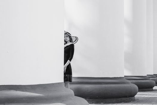 A stylish woman in a black dress and hat stands gracefully by large white columns.