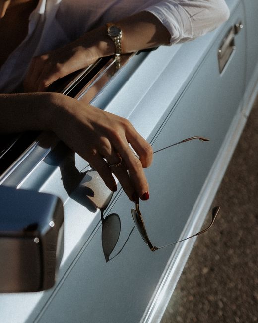 Close-up of a hand holding sunglasses out of a vintage car window on a sunny day.