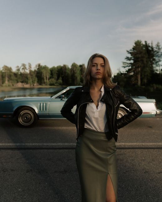 A fashion-forward woman posing confidently by a vintage car on a scenic roadside.