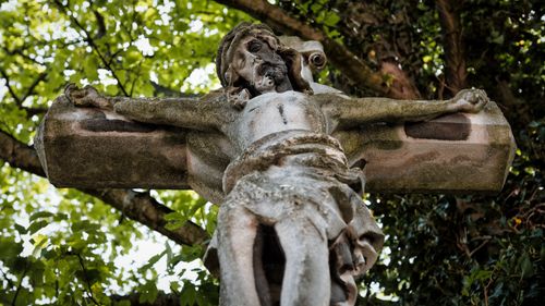 Free Low angle view of a weathered stone crucifix amidst green trees, symbolizing religious faith. Stock Photo