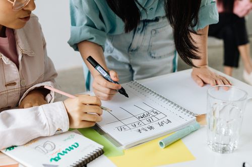 Free Two colleagues collaborate on a project, discussing and writing on a spiral notebook at their desk. Stock Photo