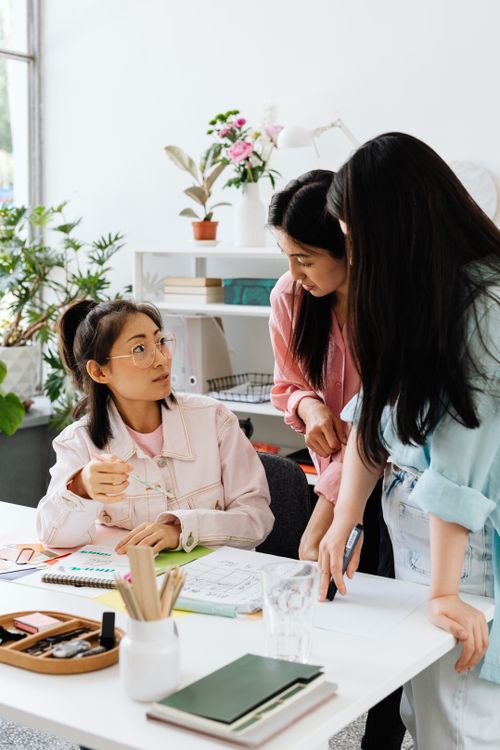 Free Three Asian women engaged in a collaborative discussion at a stylish office desk. Stock Photo