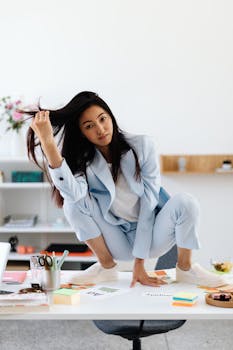 Confident woman in a blue suit positioned uniquely on a desk in a modern office.