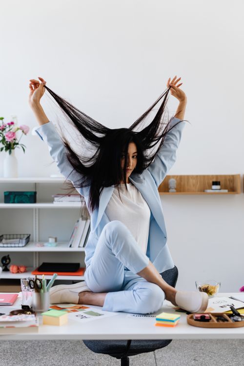 Free Fashionable woman in a blue suit playfully pulling her long hair while sitting on a desk in office setting. Stock Photo