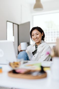 Asian woman smiling while taking a break with a ceramic mug in modern office setting.