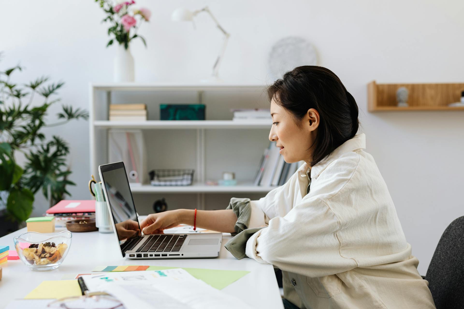 Professional Asian woman working on a laptop in a stylish, plant-decorated office setting.