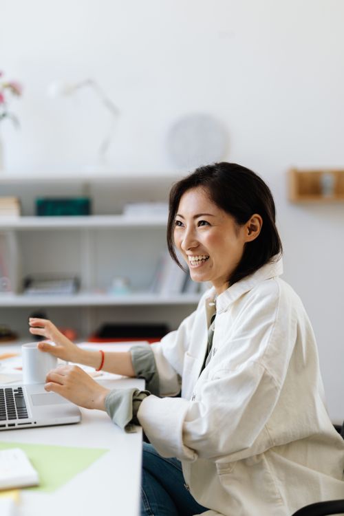 Free Asian woman in smart casual attire happily working at a desk in a bright office environment. Stock Photo