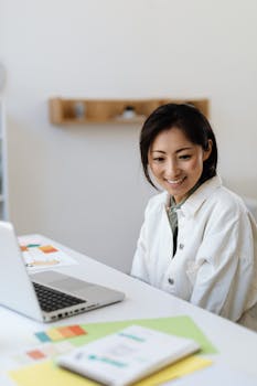 Asian woman smiling while working at desk with laptop and colorful papers in a modern office setting.