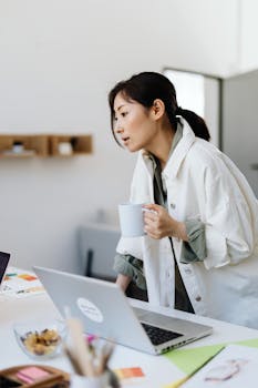 Asian woman standing in office holding a cup, working on a laptop.