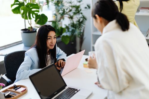 Free Businesswomen engaged in conversation while working on laptops in a modern office setting. Stock Photo