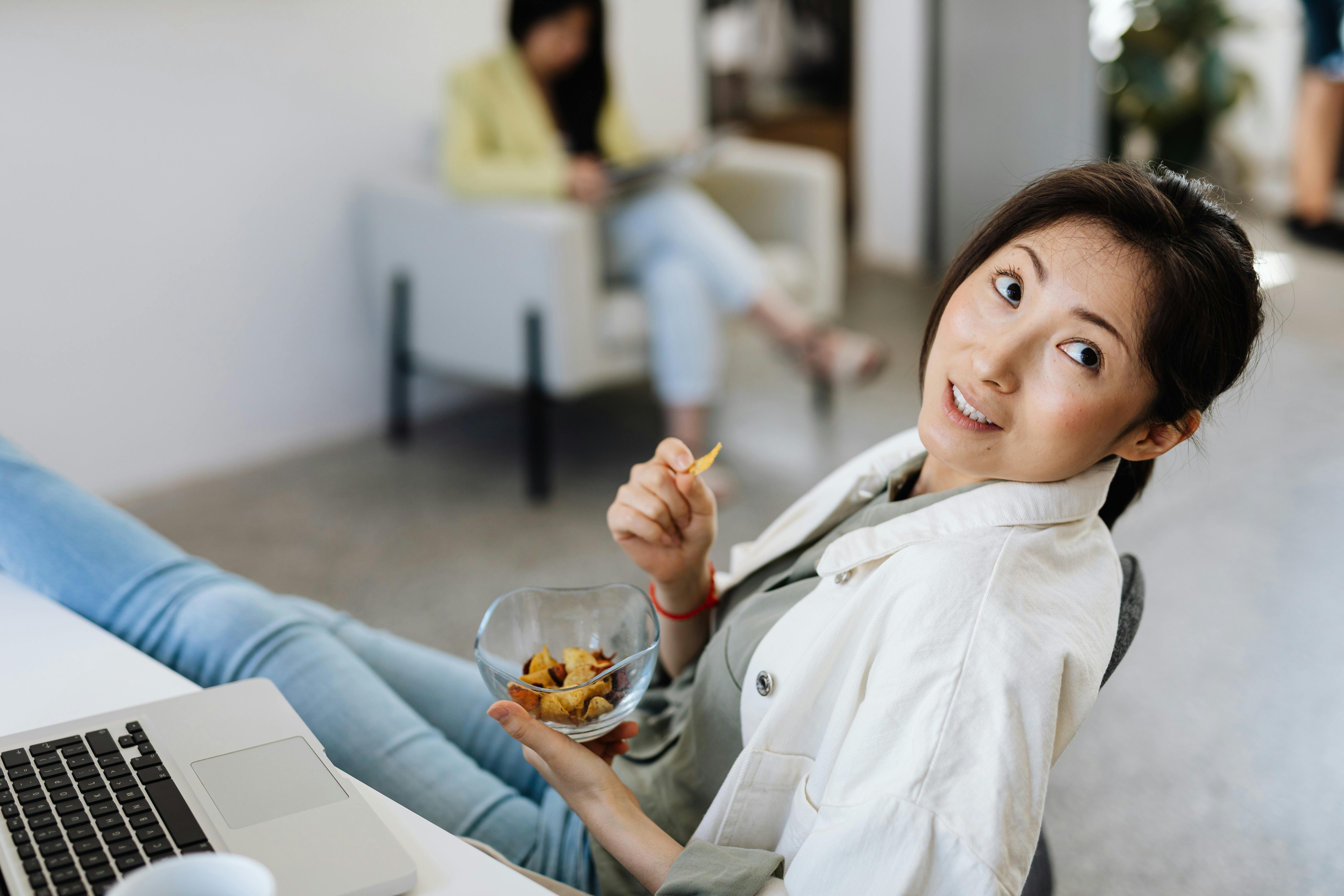 Casual scene of a woman eating chips while working on a laptop in an office setting.