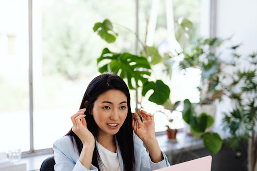 Asian woman in office, gesturing while speaking, surrounded by lush plants, near a window.