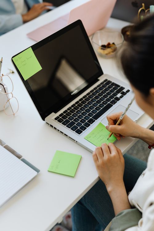 Free Overhead view of a woman writing on sticky notes using a laptop at a modern office desk. Stock Photo
