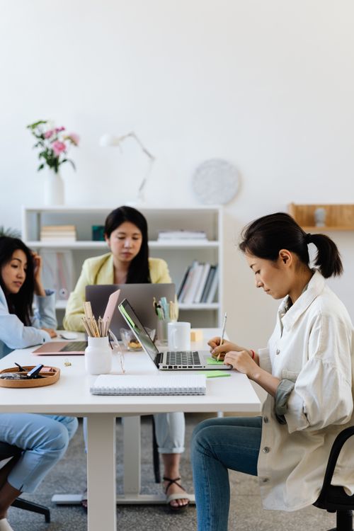 Free Three women working together at a modern office desk, focusing on laptops and notes. Stock Photo