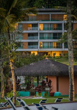 A tropical resort featuring a modern building, palm trees, and relaxing sunbeds.