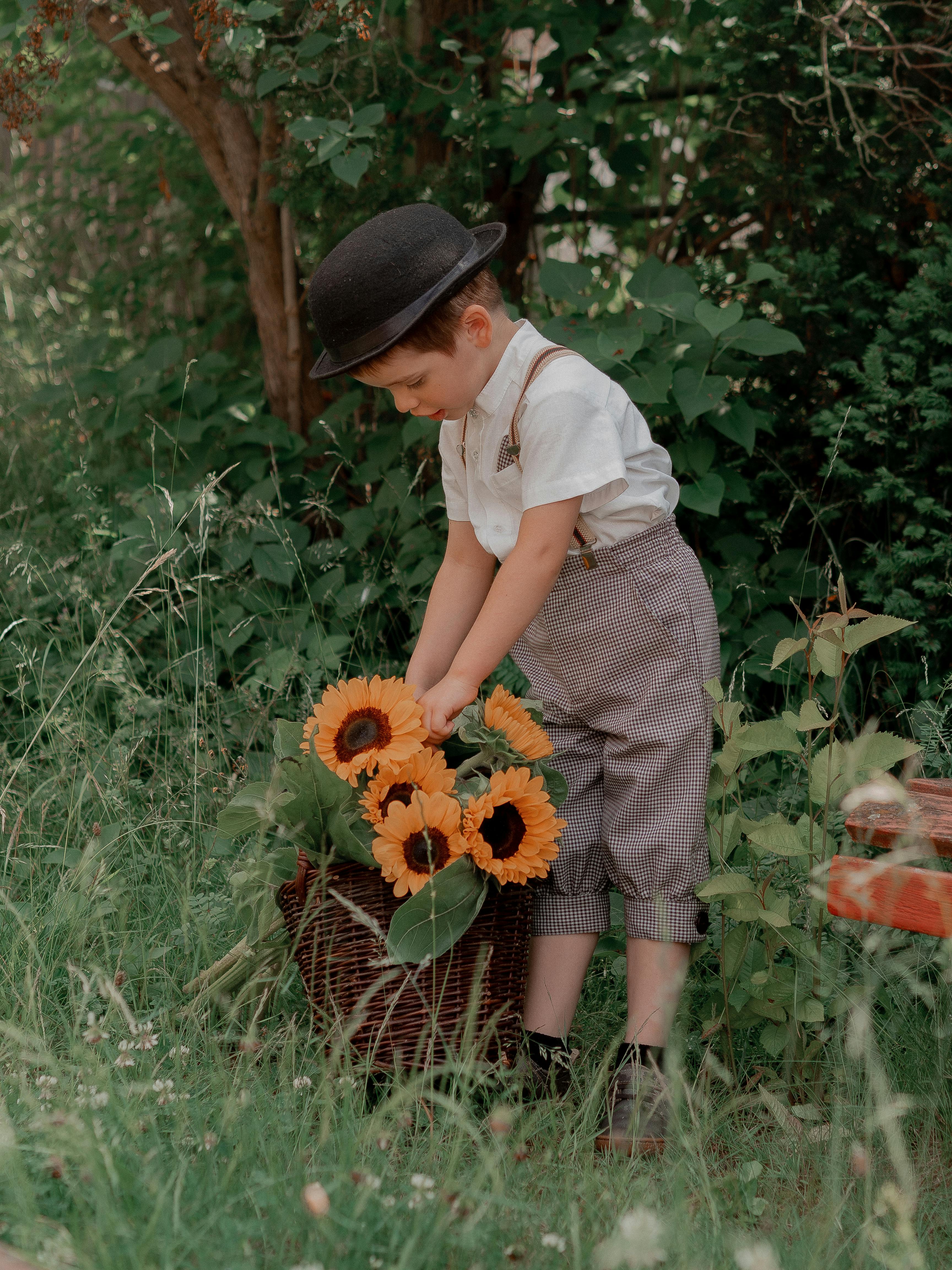 A young boy in a vintage outfit picks sunflowers from a basket outdoors in a lush garden setting.