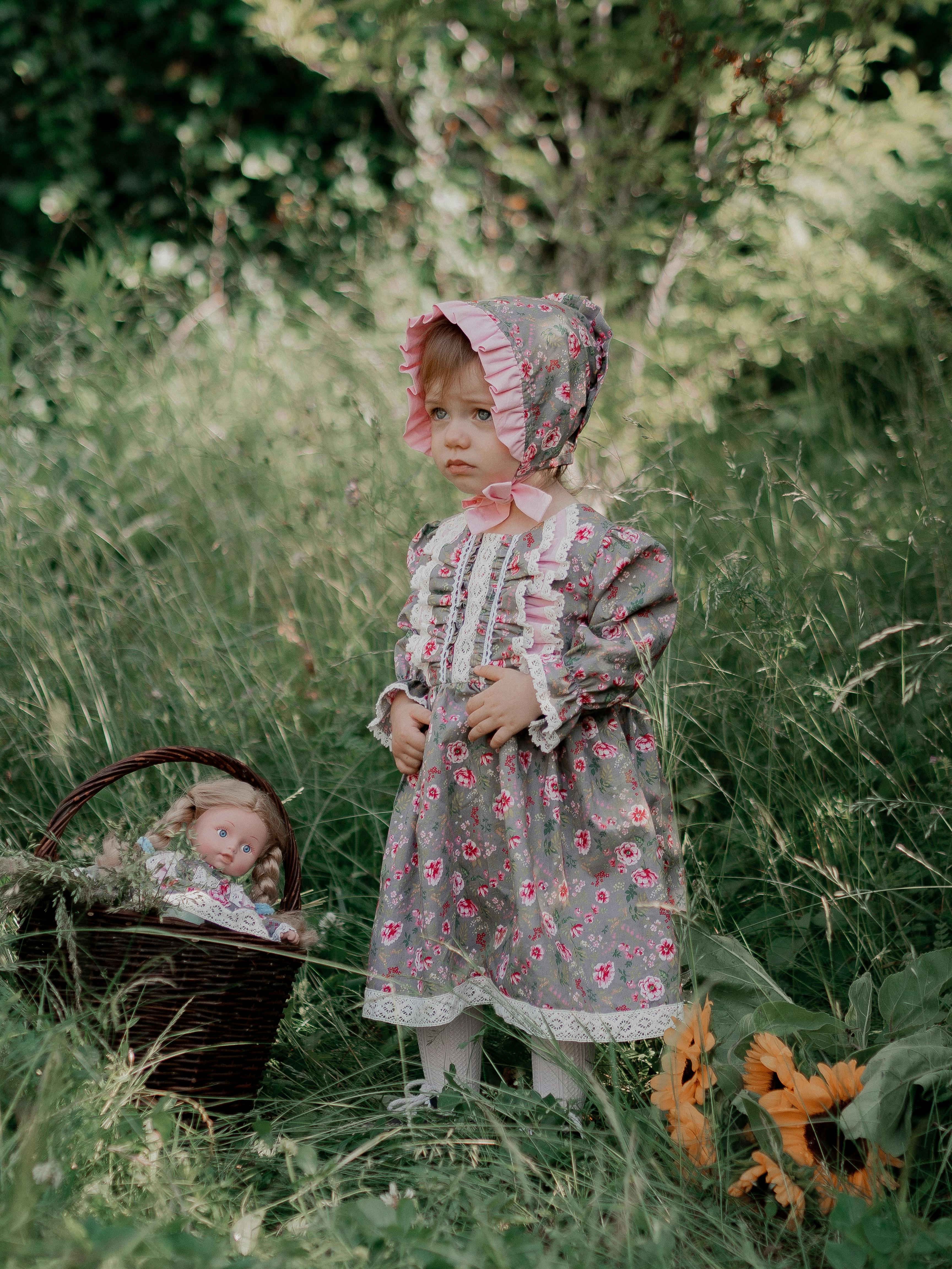 a young girl in floral dress standing on grass field near the woven basket with doll