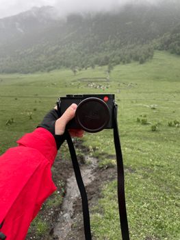A hand holding a camera in a lush green meadow with hills in the background, perfect for nature photography.
