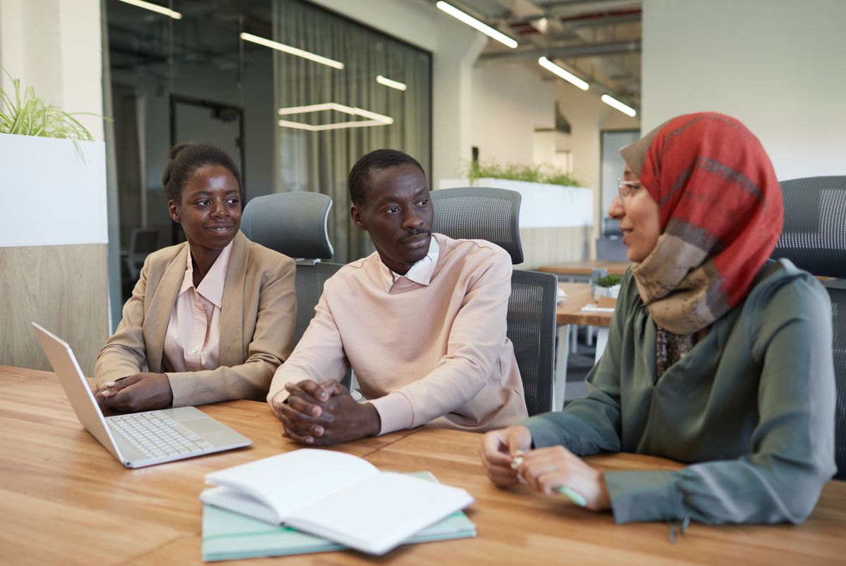 Three colleagues talk through their work at a meeting table with laptops and notebooks