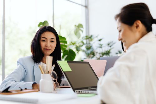 Free Two Asian women working in a bright, modern office setting with laptops and office supplies. Stock Photo