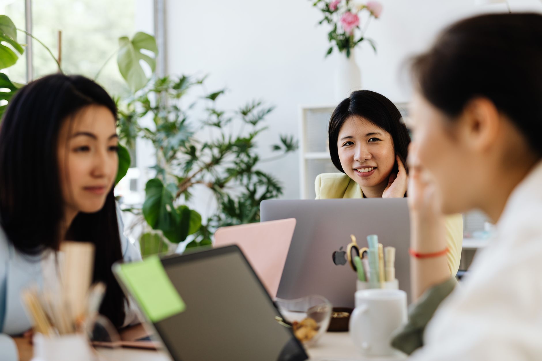 Three women in a modern office setting discussing work with laptops.