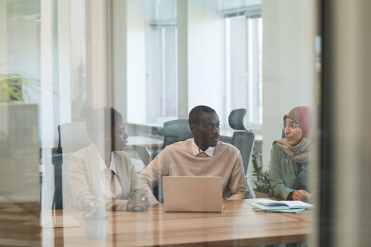 Three professionals in a modern office setting engaged in a discussion around a laptop, showcasing teamwork and collaboration.