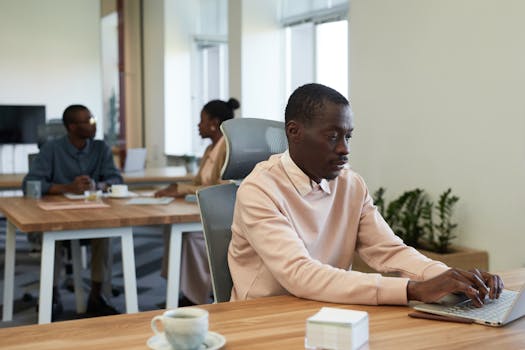 Black man concentrating while working on a laptop at a modern office, collaborating with colleagues.